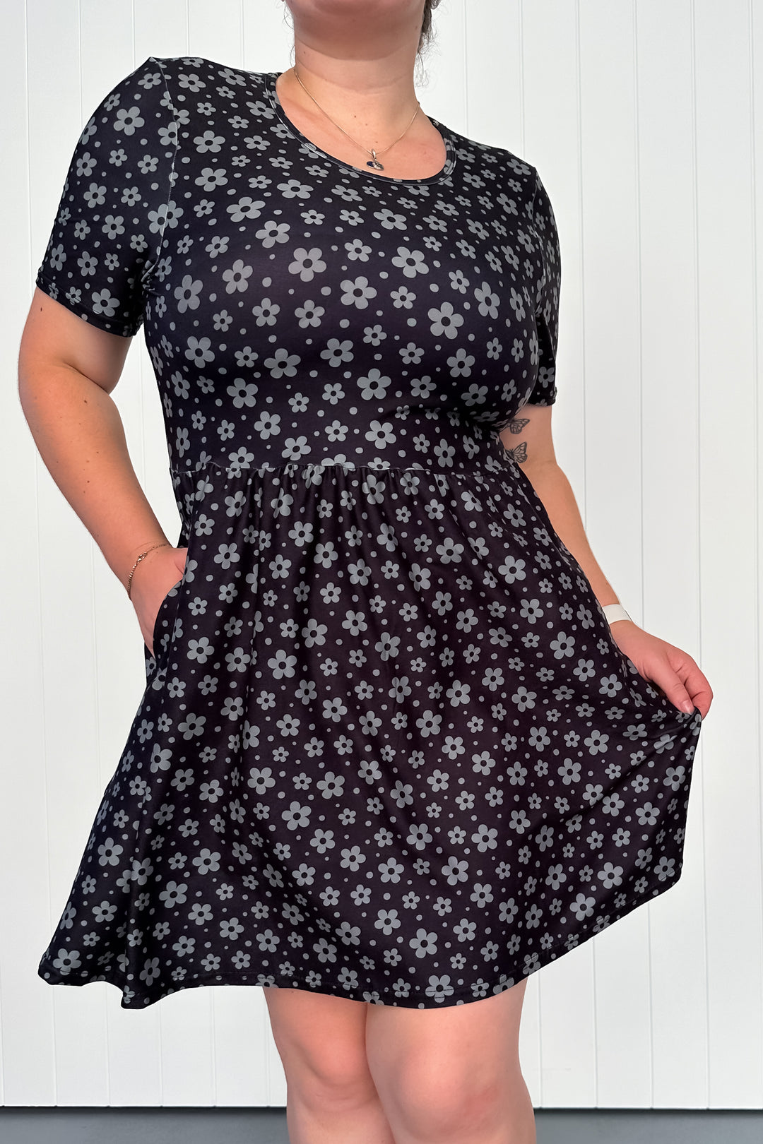 A model wearing a short-sleeved mini dress with a deep-black background featuring a delicate daisy motif. The dress has deep side pockets and appears to have a vintage-inspired shape.