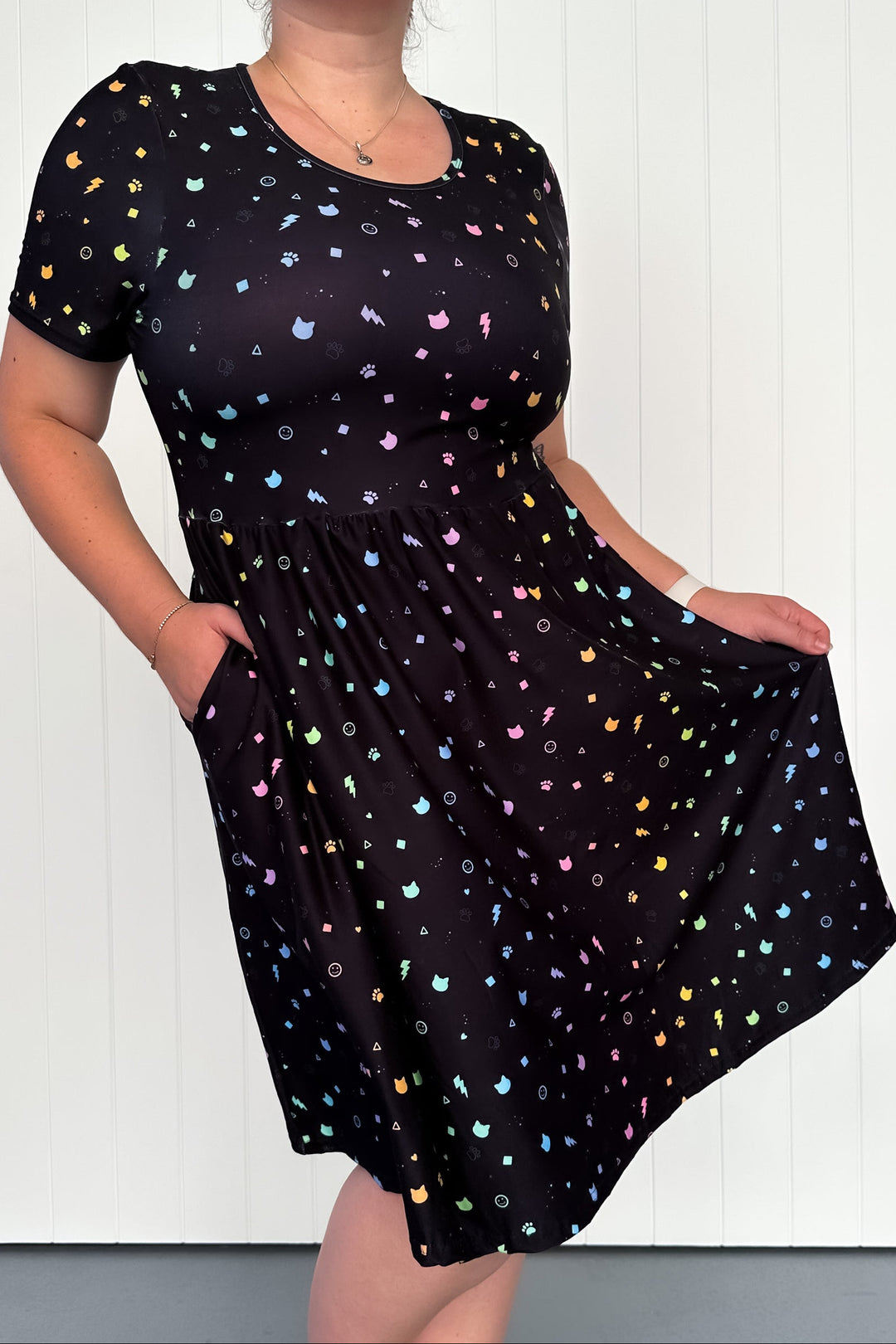 A knee-length black dress with a pattern of colorful cat faces, paw prints, and geometric shapes. The dress has short sleeves and side pockets.