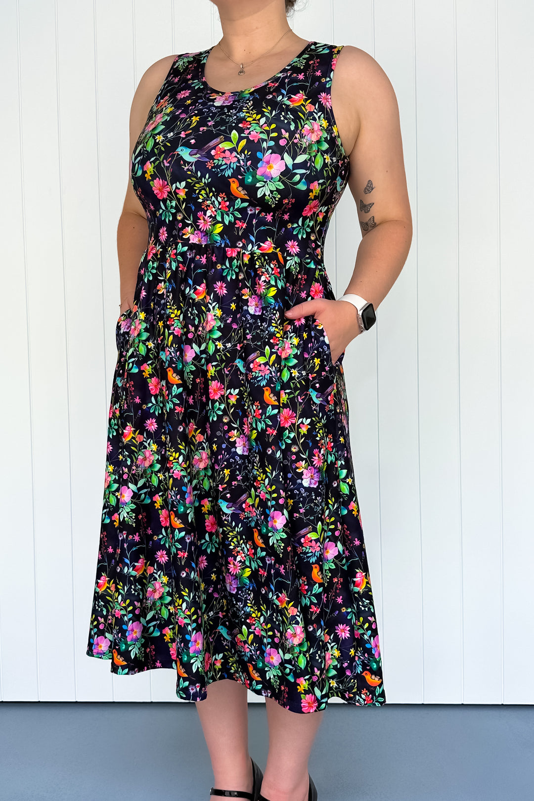 A sleeveless midi dress with a floral pattern featuring colors such as pink, orange, and green on a dark background and colourful birds. The dress has a square neckline, and deep side pockets.
