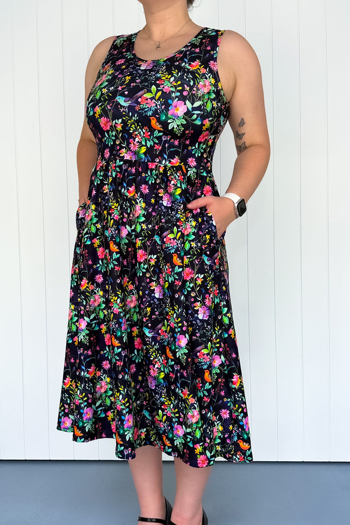 A sleeveless midi dress with a floral pattern featuring colors such as pink, orange, and green on a dark background and colourful birds. The dress has a square neckline, and deep side pockets.
