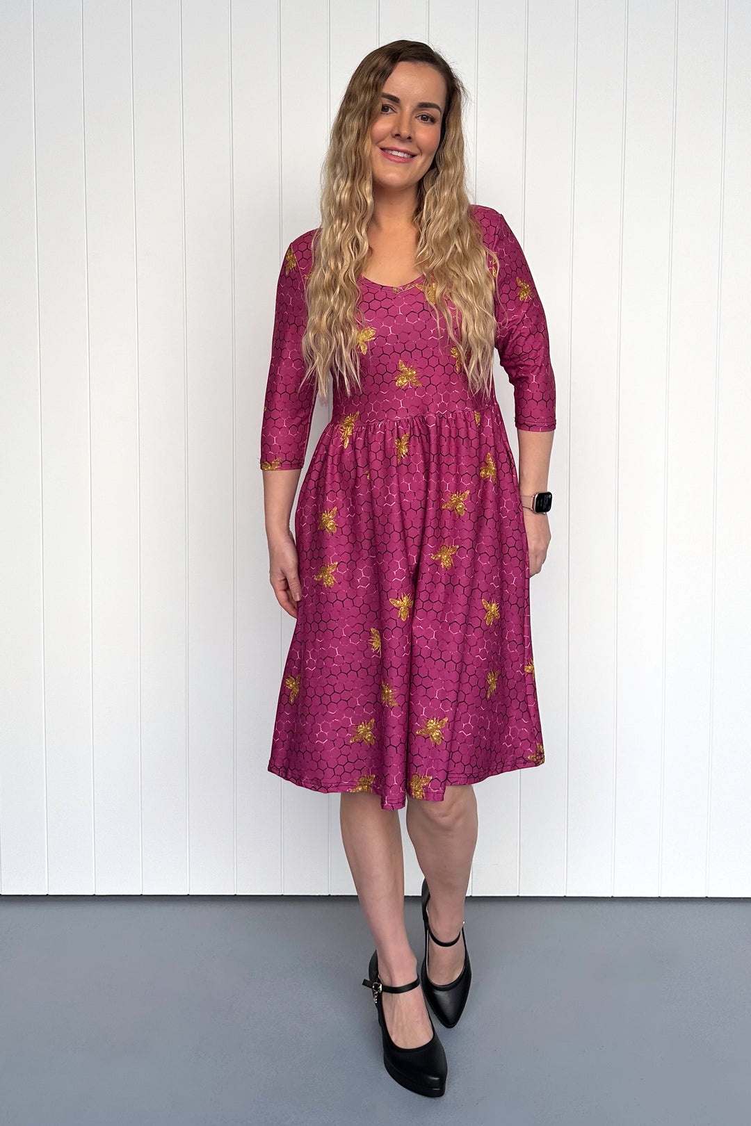 Woman wearing a magenta dress with golden bee patterns against a white wall.