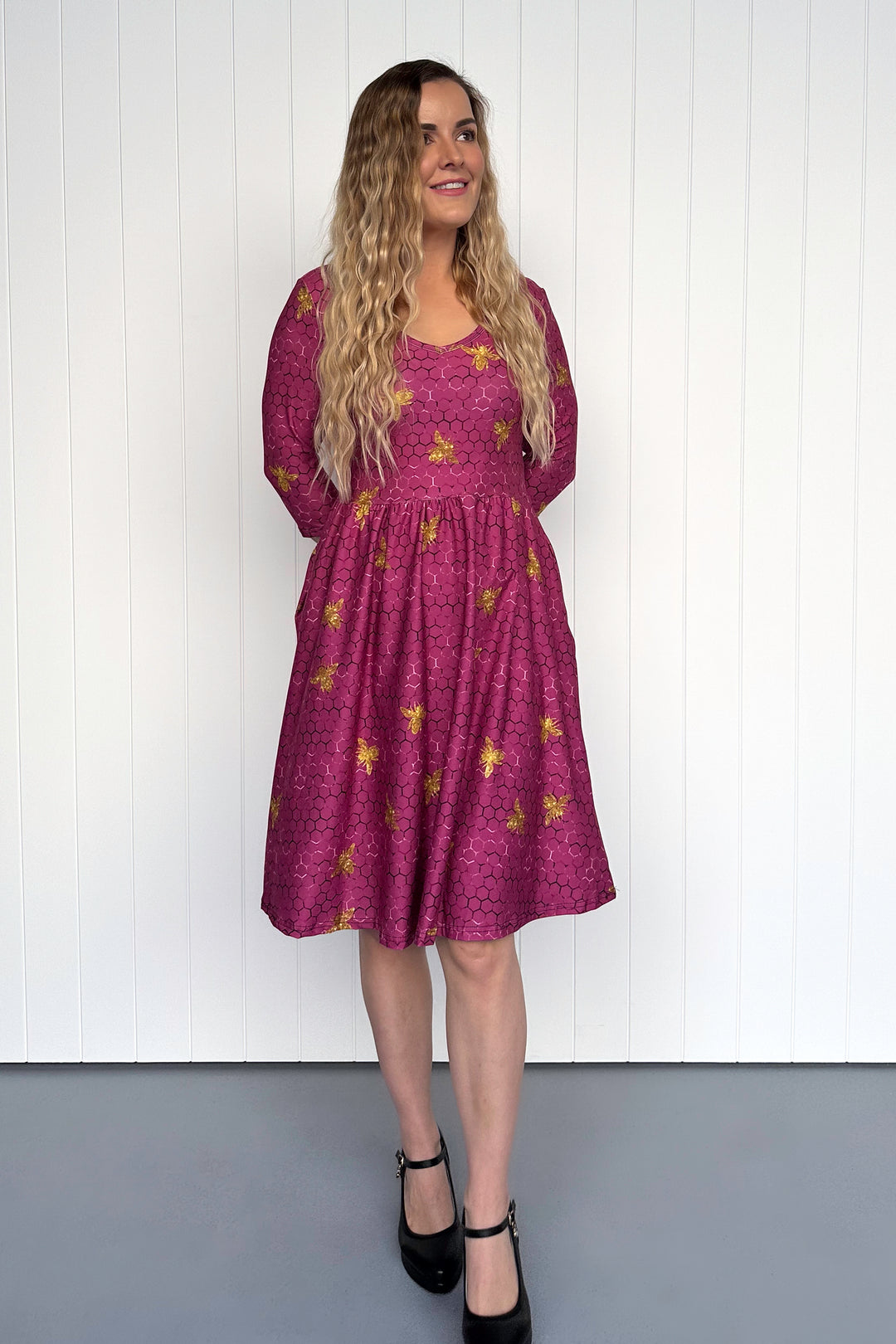 Woman wearing a magenta dress with golden bee patterns against a white wall.