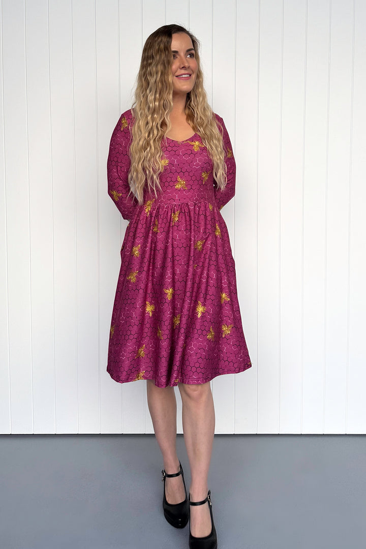 Woman wearing a magenta dress with golden bee patterns against a white wall.