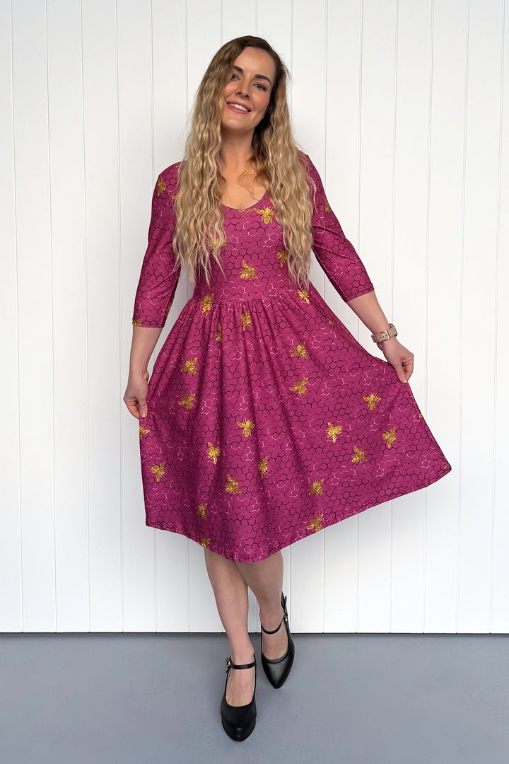 Woman wearing a pink dress with golden bee patterns against a white wall.