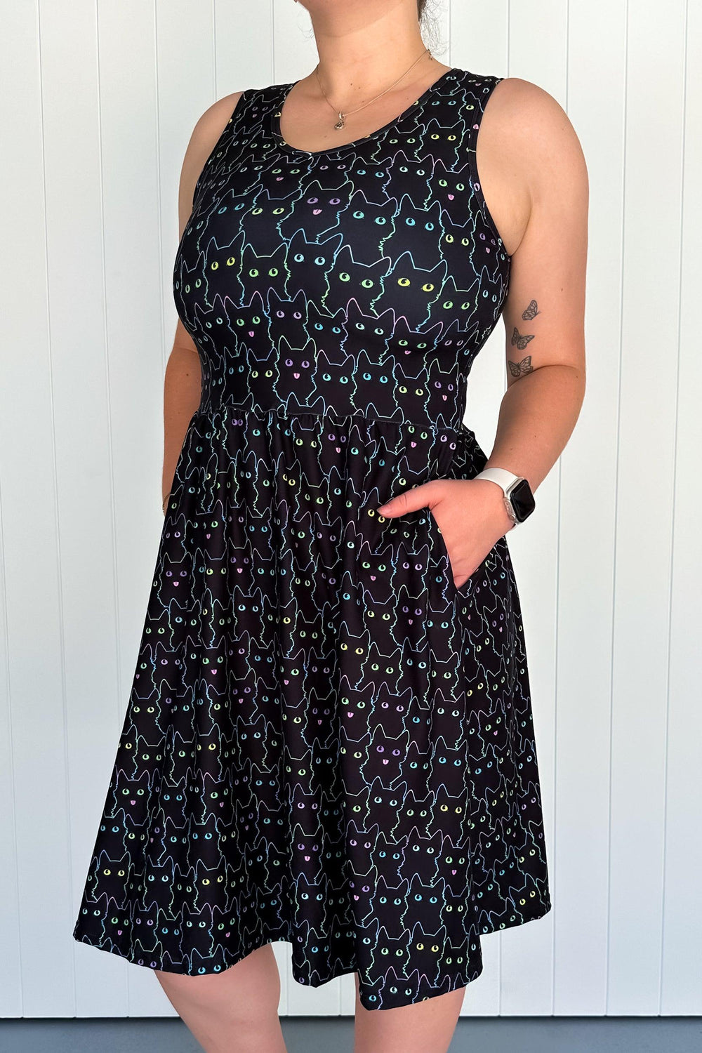 A sleeveless knee-length black dress with a pattern of black feline silhouettes and multicolored reflective eyes, worn by a model showing the side pockets.
