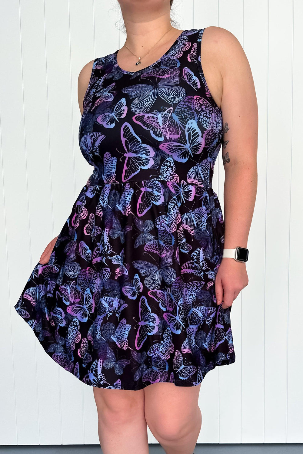A sleeveless mini dress with a butterfly pattern in jewel tones on a black background, featuring a round neckline and side pockets.
