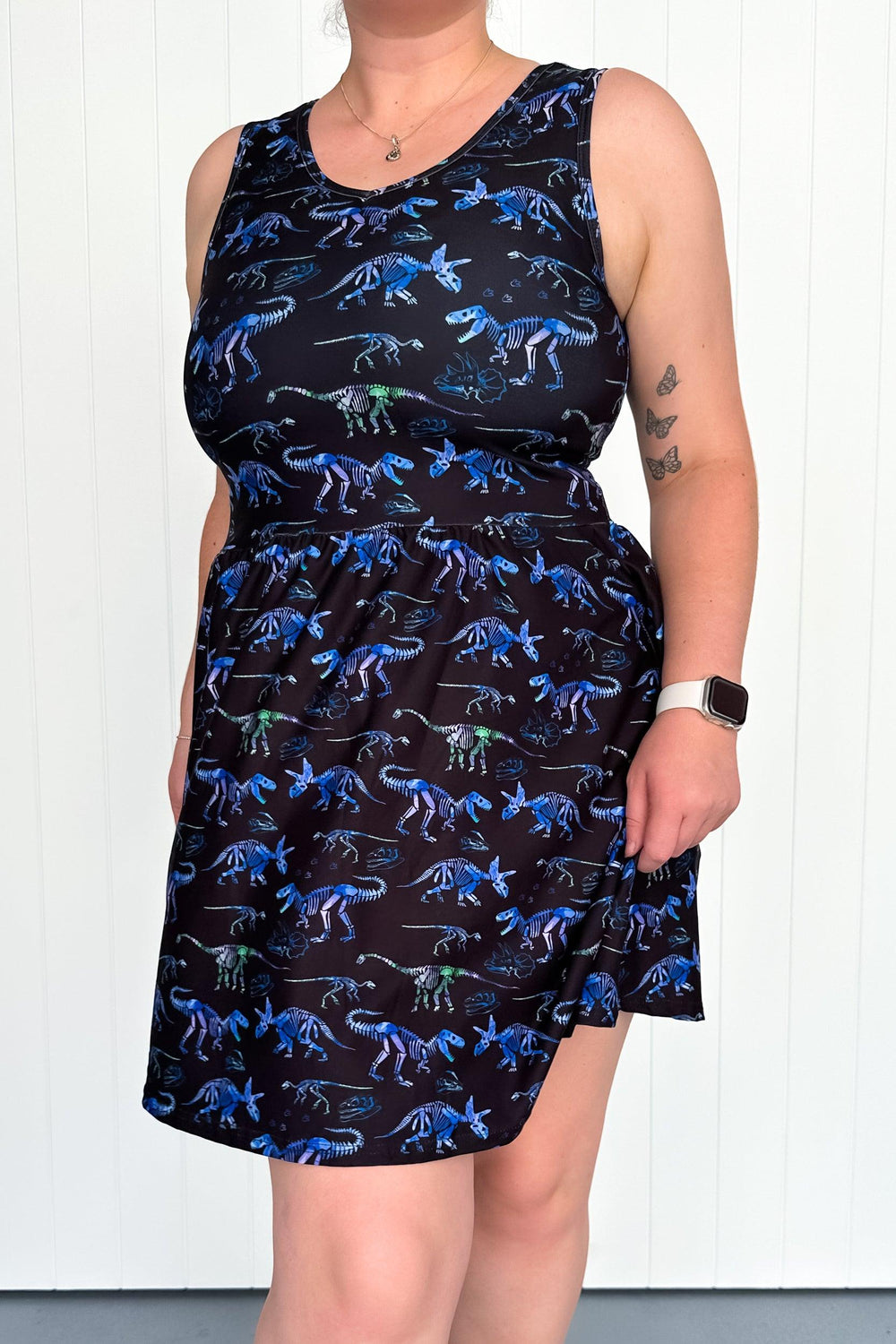 A sleeveless mini dress with a black background covered in blue and green dinosaur patterns, featuring side pockets.