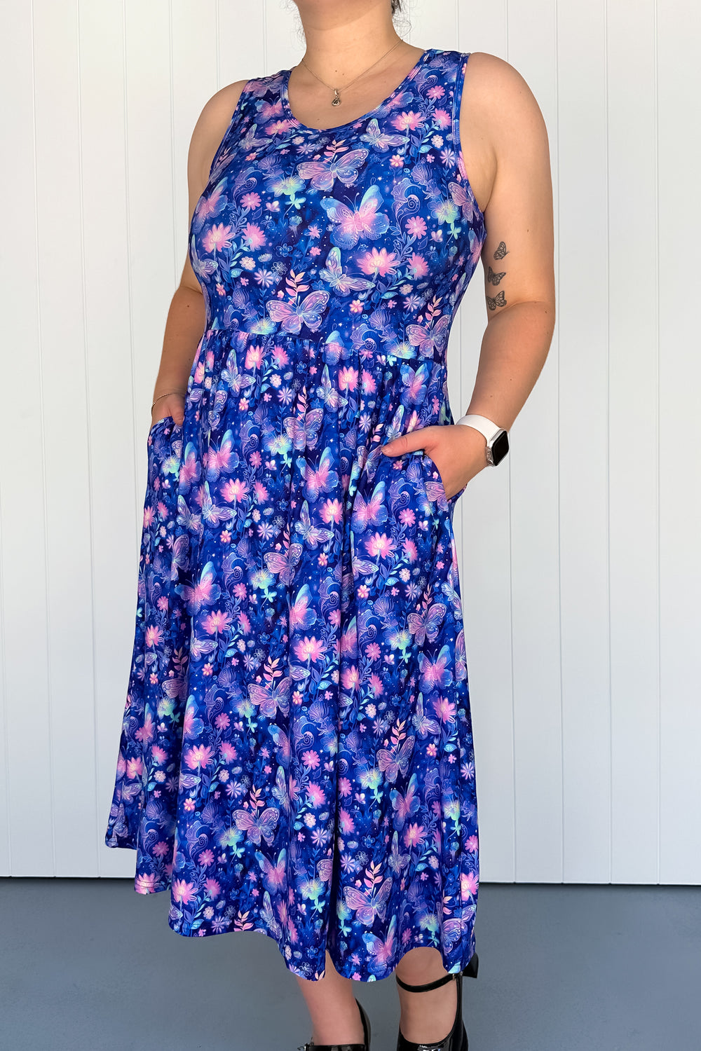 A sleeveless midi dress with a floral pattern featuring soft pink butterflies and tiny florals on a deep twilight blue base. The dress has a round neckline and deep side pockets.